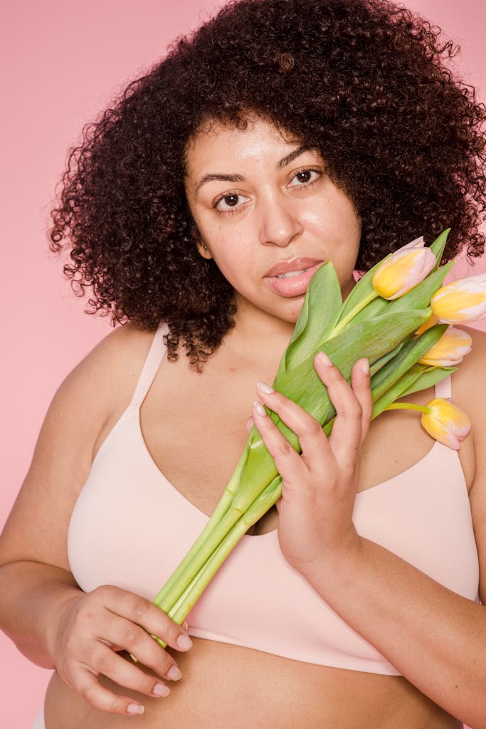 Plus size African American female model wearing beige lingerie looking at camera on pink background with fresh tulips in hands
Self-trust