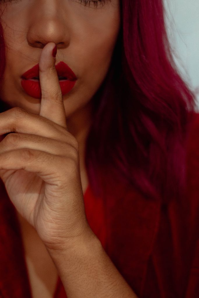Close-up of a woman with pink hair, red lipstick, making a shushing gesture.