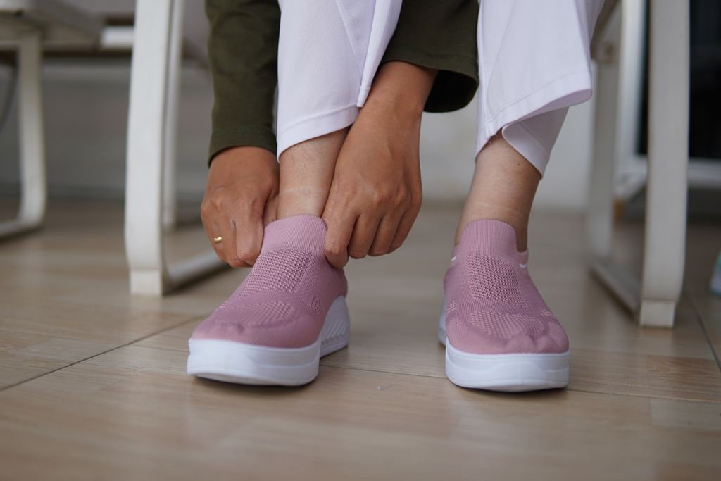 Close-up of someone putting on pink slip-on sneakers while seated indoors on wooden flooring.